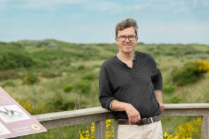 Portrait von Herr Vodde auf einem Bohlenweg. Im Hintergrund sind Dünen zu sehen.