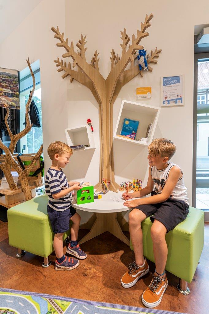 Das Bild zeigt eine Kinderecke in der Touristinformation in Wolfenbüttel. Es ist ein Tisch mit zwei Hockern zu sehen, ein Straßenteppich und verschiedene weitere Spielmöglichkeiten. Aus dem Tisch ragt ein hölzerner Baum, die ganze Dekoration ist sehr lebendig.